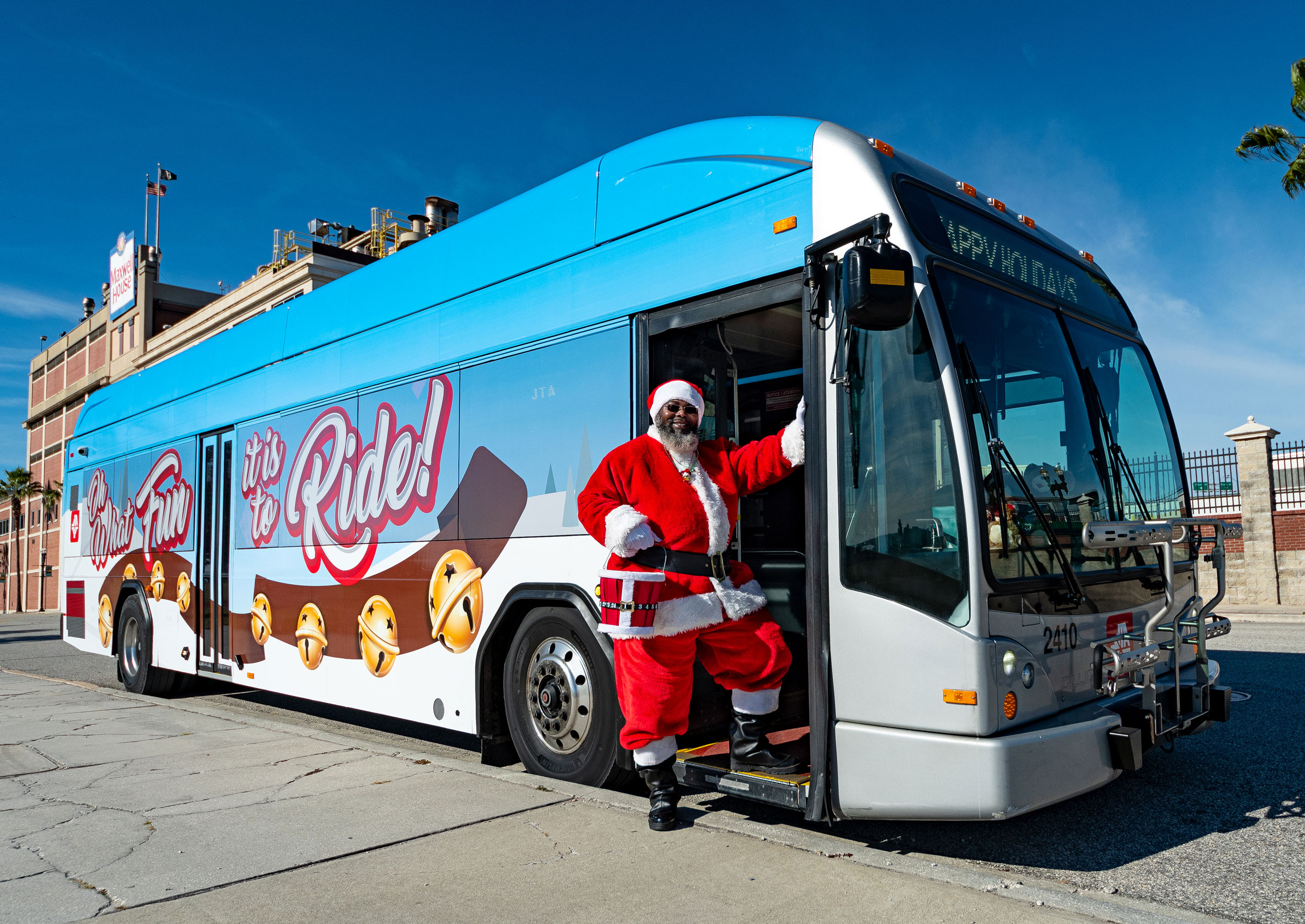 A Jacksonville Transportation Authority bus is parked along on a city street with winter holiday graphics shown on the side. A bus driver dressed as Santa is standing beside the bus.
